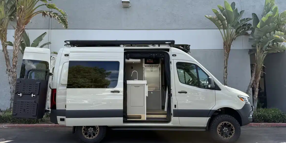 Ein weißer Campervan mit geöffneter Seitentür, sichtbarer Küche und robustem Offroad-Design, steht vor einer grauen Wand zwischen Palmen.