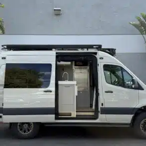 Ein weißer Campervan mit geöffneter Seitentür, sichtbarer Küche und robustem Offroad-Design, steht vor einer grauen Wand zwischen Palmen.
