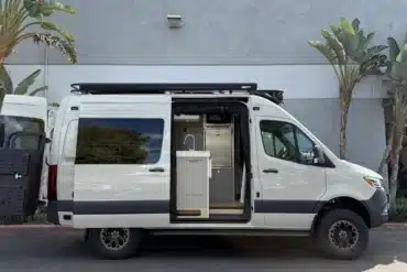 Ein weißer Campervan mit geöffneter Seitentür, sichtbarer Küche und robustem Offroad-Design, steht vor einer grauen Wand zwischen Palmen.