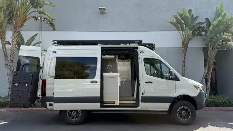 Ein weißer Campervan mit geöffneter Seitentür, sichtbarer Küche und robustem Offroad-Design, steht vor einer grauen Wand zwischen Palmen.