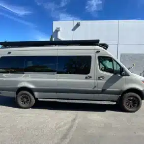 Seitenansicht eines ausgebauten Mercedes-Benz Sprinter 170 4x4 Campervans mit langem Radstand und Offroad-Bereifung vor einem modernen Gebäude.