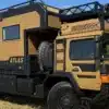 Das MAN HX60 Atlas Expeditionsmobil, Modelljahr 2024, auf einer Wiese, in braun-schwarzer Lackierung mit markantem Offroad-Ausbau, großen Reifen und abnehmbarer Wohnkabine.