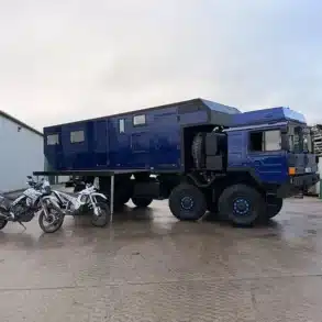 MAN HX77 Expeditionsmobil mit großem Wohnaufbau und Motorrädern auf einem Industriegelände