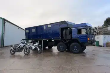 MAN HX77 Expeditionsmobil mit großem Wohnaufbau und Motorrädern auf einem Industriegelände