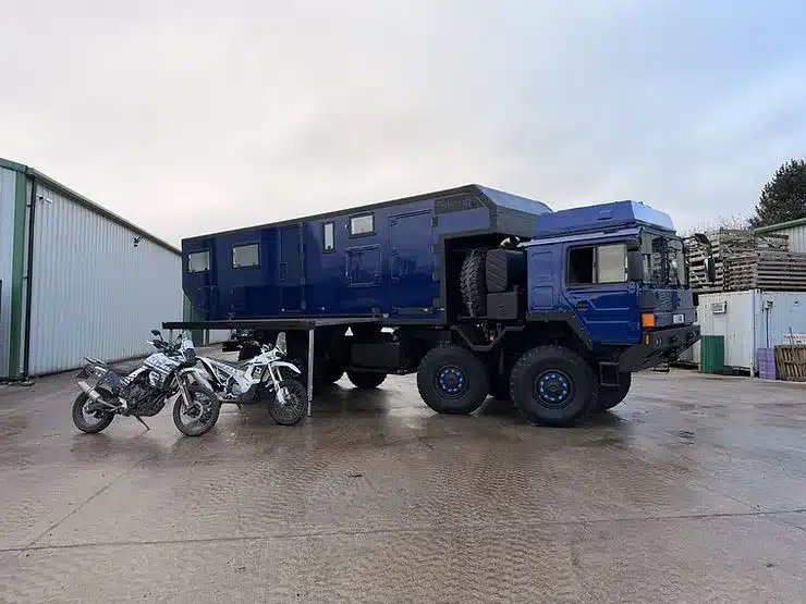 MAN HX77 Expeditionsmobil mit großem Wohnaufbau und Motorrädern auf einem Industriegelände
