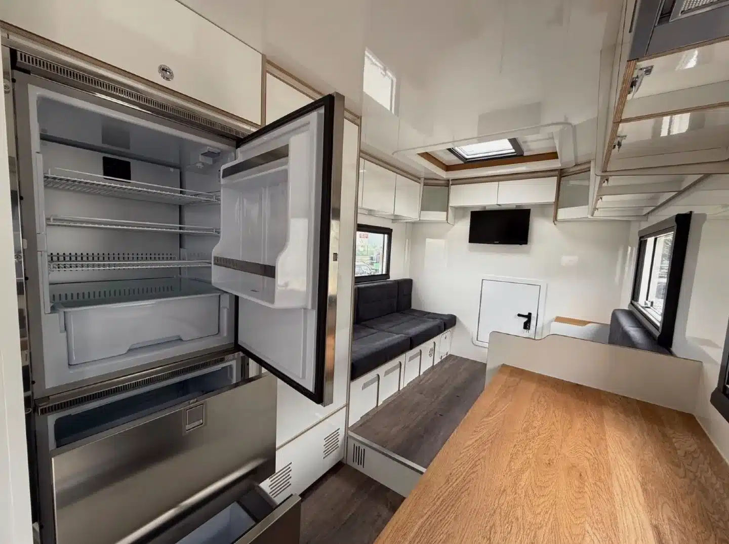 Moderner Innenraum eines Expeditionsmobils mit geöffneter Kühl-/Gefrierkombination, Couch, Stauraum und TV.