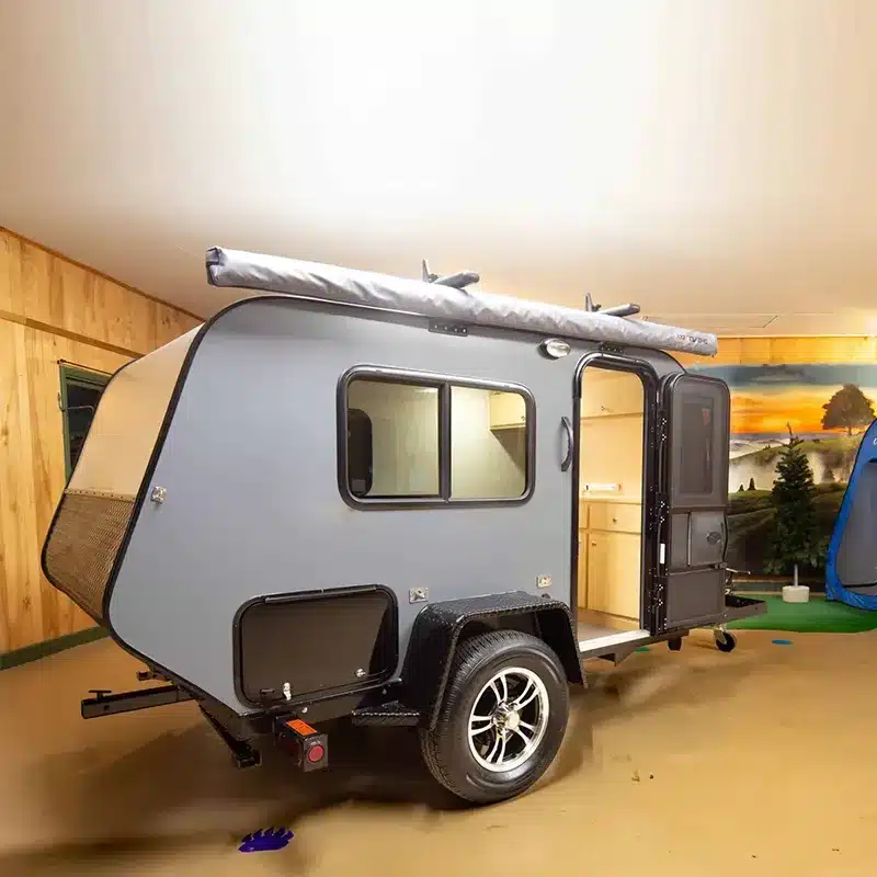 Kompakter Mini 2D Offroad-Camper-Trailer mit robustem Chassis und Markise, Innenraum im Hintergrund sichtbar.