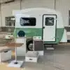 MOOI Mini Teardrop Camper Trailer mit aufgeklappter Küche, Sitzhockern und Tisch vor einer Industriehalle