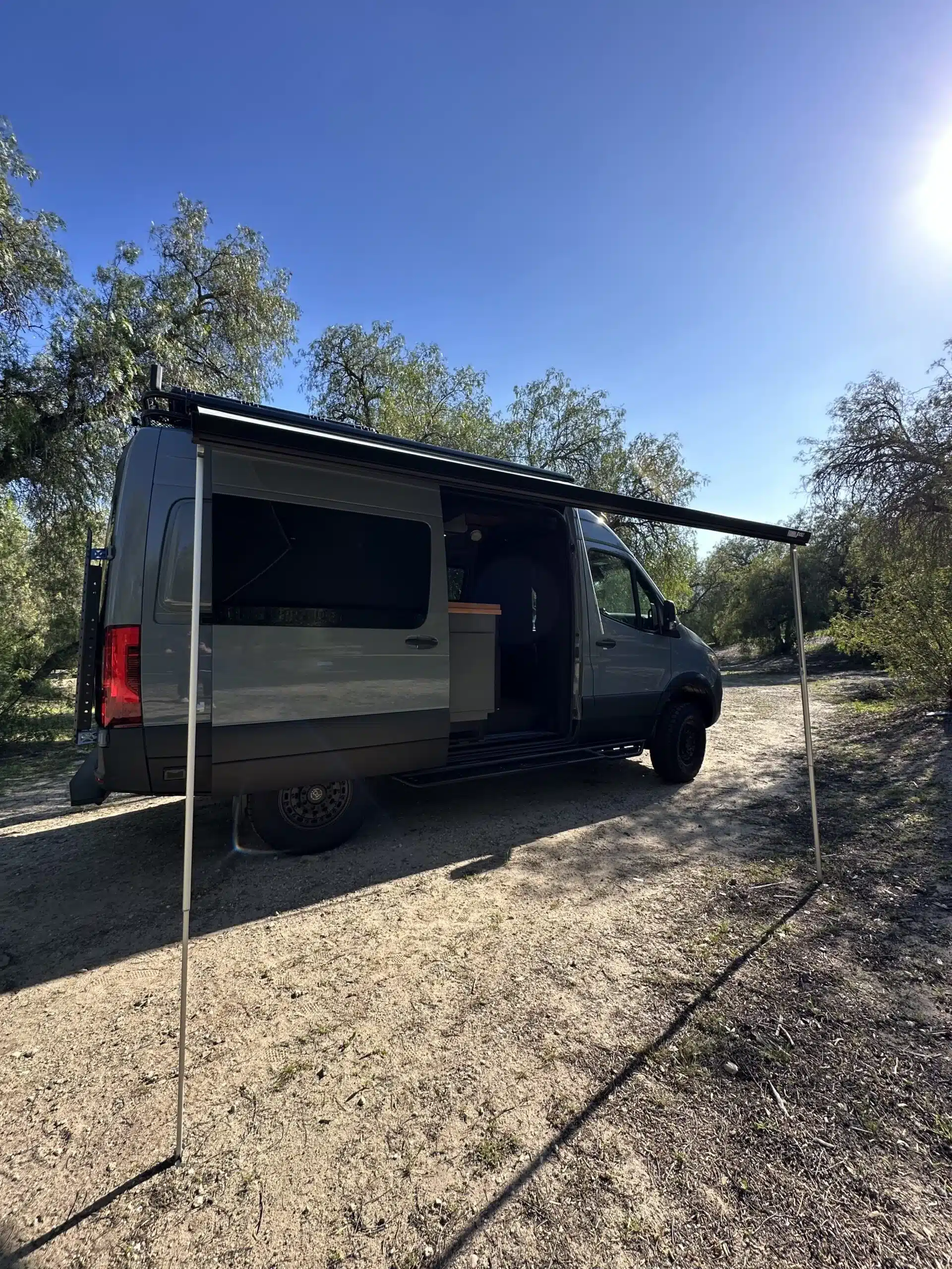 Außenansicht des Nestor Campervans, Mercedes Sprinter 144 AWD, mit geöffneter Schiebetür und ausgefahrener Markise, auf einem naturbelassenen Stellplatz.