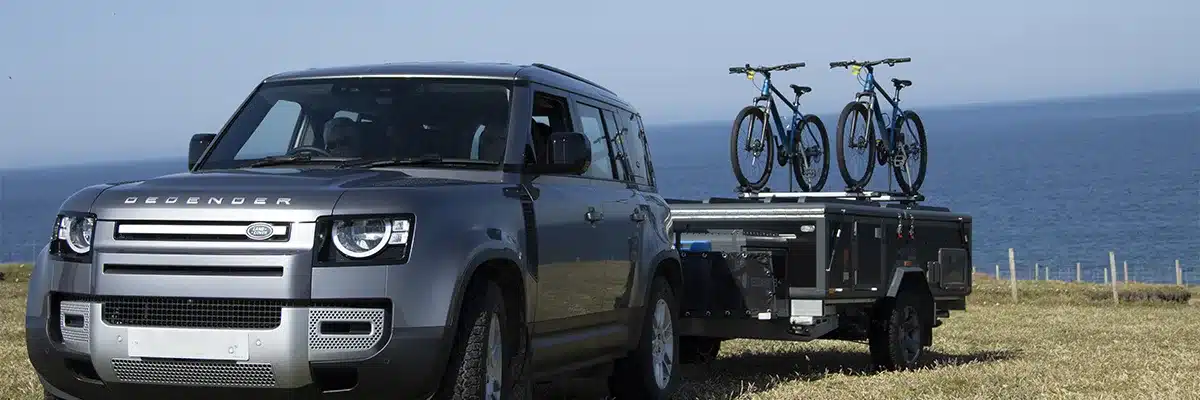 Off-Road Air OPUS Offroad-Faltcaravan, gezogen von einem Land Rover Defender mit zwei Fahrrädern auf dem Dach, am Küstenstreifen.