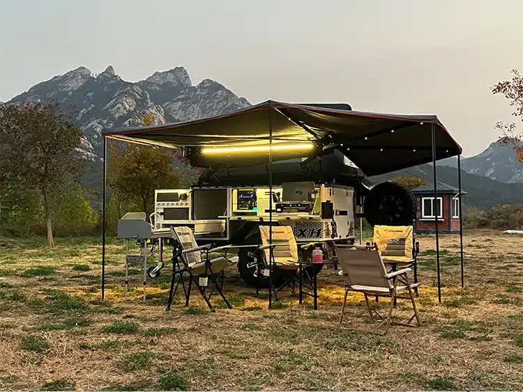 Patriot X1H Travel Trailer mit Markise, Outdoor-Küche und Campingmöbeln auf einer Wiese vor Bergen.