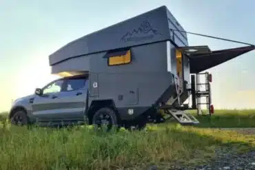Pickup mit aufgesetzter Wohnkabine samt elektrischem Parallelhubdach, geparkt auf einer Wiese bei Sonnenuntergang.