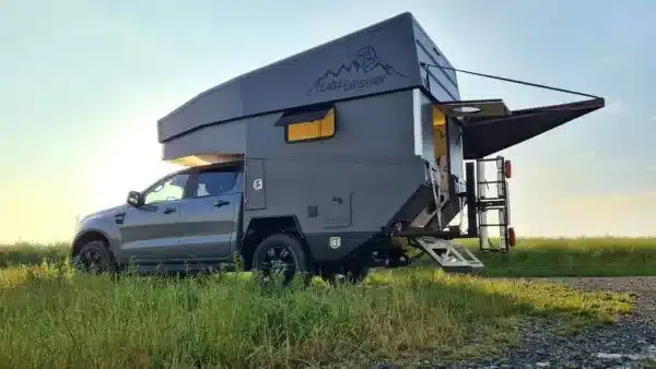 Pickup mit aufgesetzter Wohnkabine samt elektrischem Parallelhubdach, geparkt auf einer Wiese bei Sonnenuntergang.