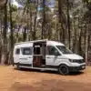 Weißer Reviva Camper Van von Hotomobil steht mit geöffneter Tür in einem lichten Wald auf sandigem Boden.