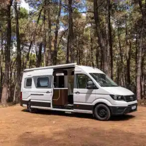 Weißer Reviva Camper Van von Hotomobil steht mit geöffneter Tür in einem lichten Wald auf sandigem Boden.