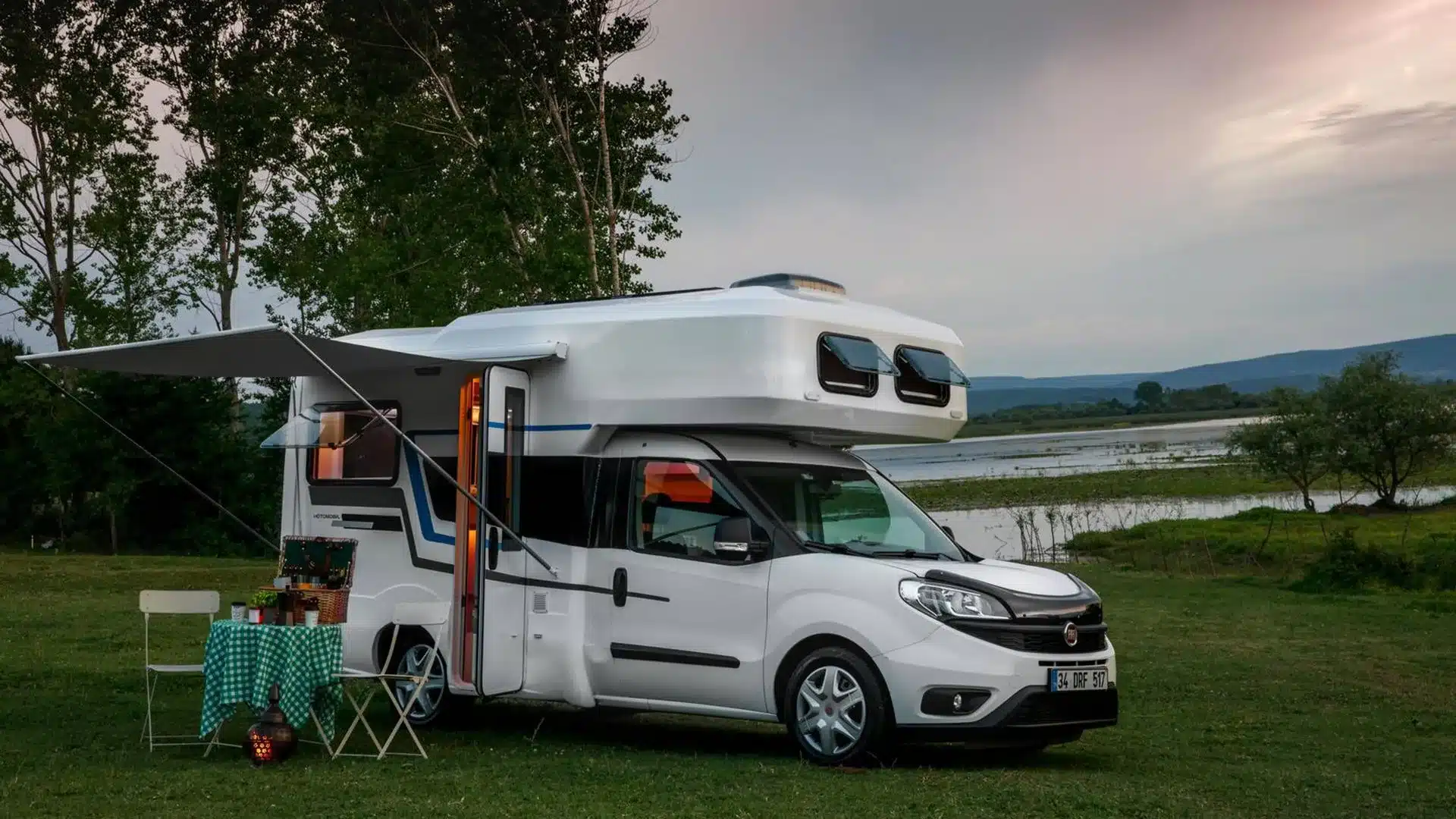 Ronin XL Motorhome mit Markise und Picknick auf Wiese am Seeufer bei Sonnenuntergang.