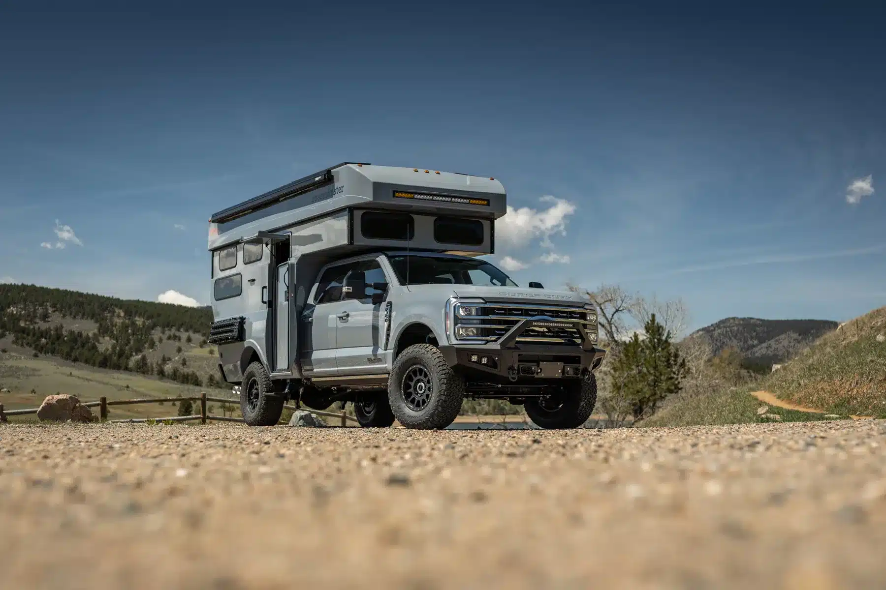 Rossmönster Baja Truckcamper mit aufstellbarem Hartschalendach auf US-Pick-up in offener Landschaft vor bewaldeten Hügeln
