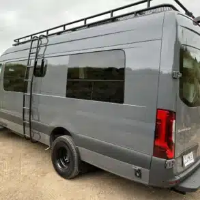 Seitenansicht des ausgebauten Mercedes Sprinter Vans 'Stitch' mit Dachträger und Leiter auf unbefestigtem Weg.