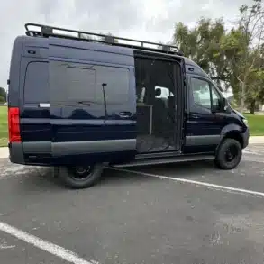 Blauer Adventure-Van The Blue Wanderer auf Parkplatz, Schiebetür geöffnet, Insektenschutz sichtbar.