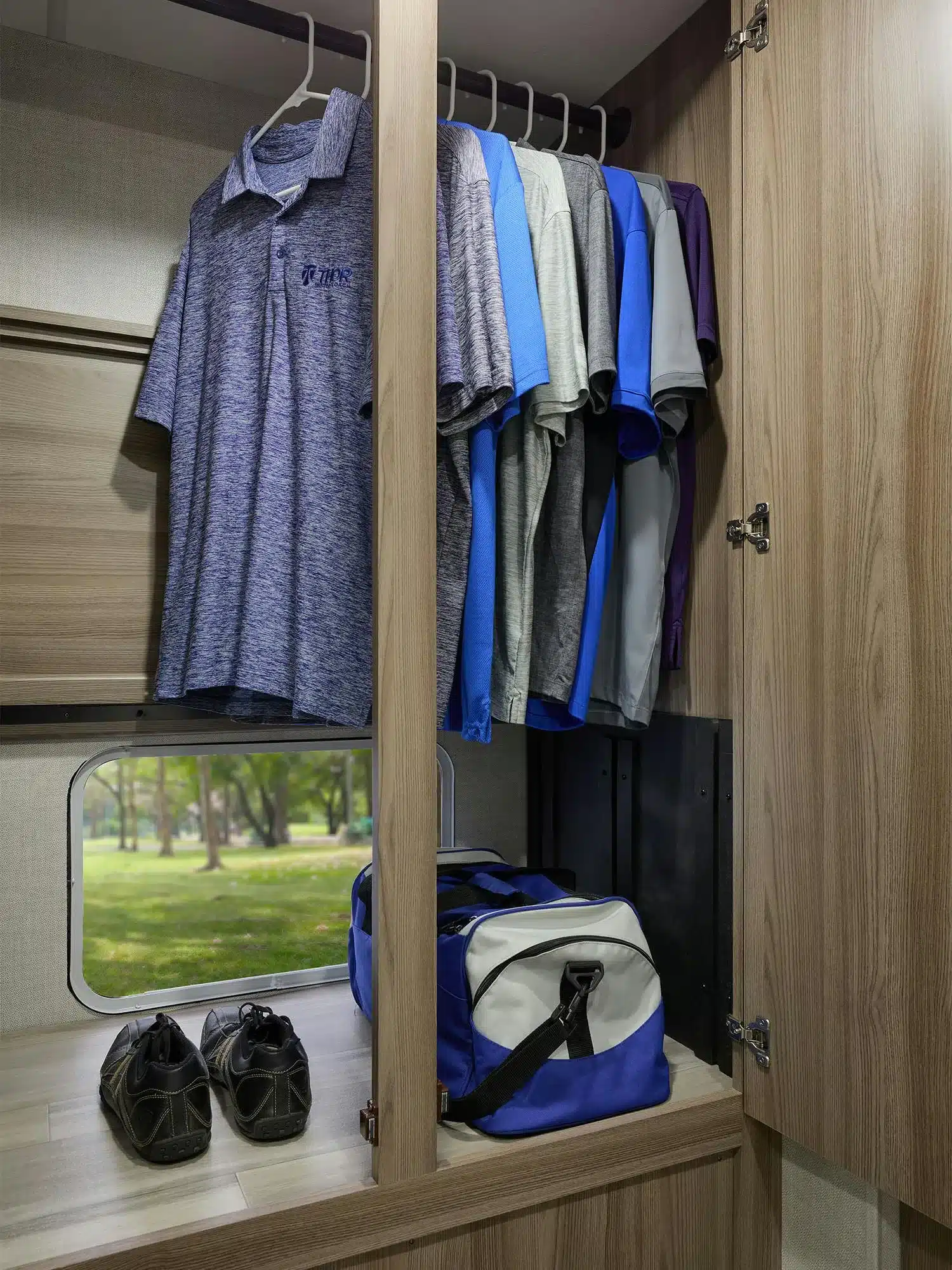 Kleiderschrank mit aufgehängter Kleidung, Sportschuhen und Tasche im Thor Gemini AWD 24KB, mit Blick aus dem Fenster ins Grüne.
