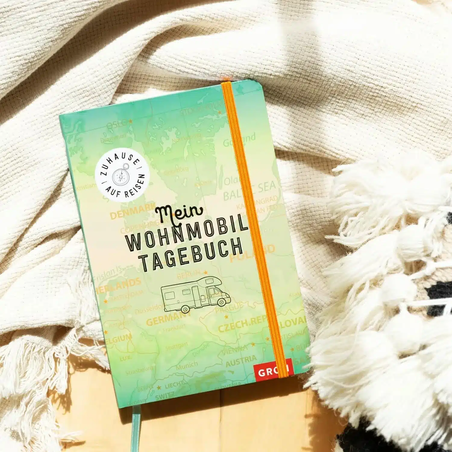 Wohnmobil-Tagebuch mit grüner Europakarte als Umschlag auf heller Decke liegend, orange Gummiband, Wohnmobil- und Kompassgrafik
