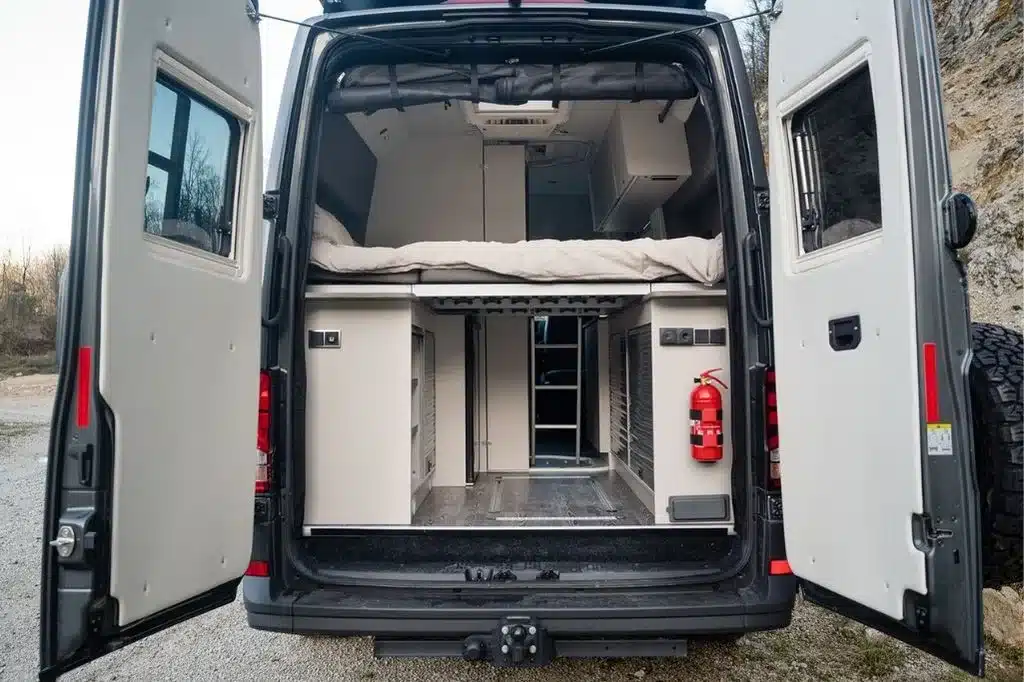 Blick in den geöffneten Heckbereich eines Aquila Adventure Creston Crafter 4x4, sichtbar sind Bett, Stauraum und Feuerlöscher.