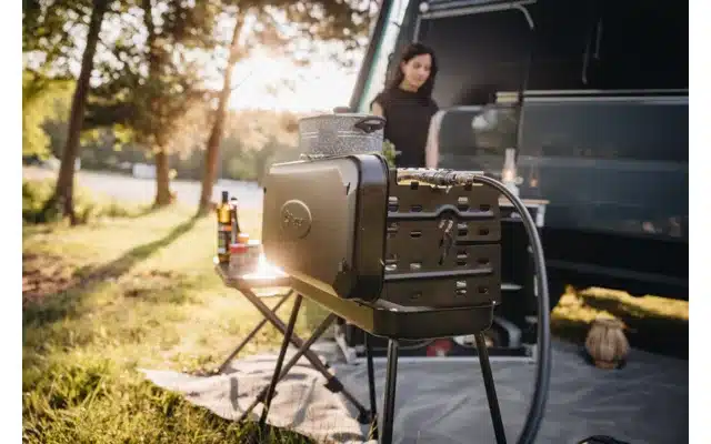 Berger 3-Flammen Koffergasgrill Schwarz auf einem Campingplatz, angeschlossen und einsatzbereit vor einem Campervan, mit Kochzubehör auf dem Grill.