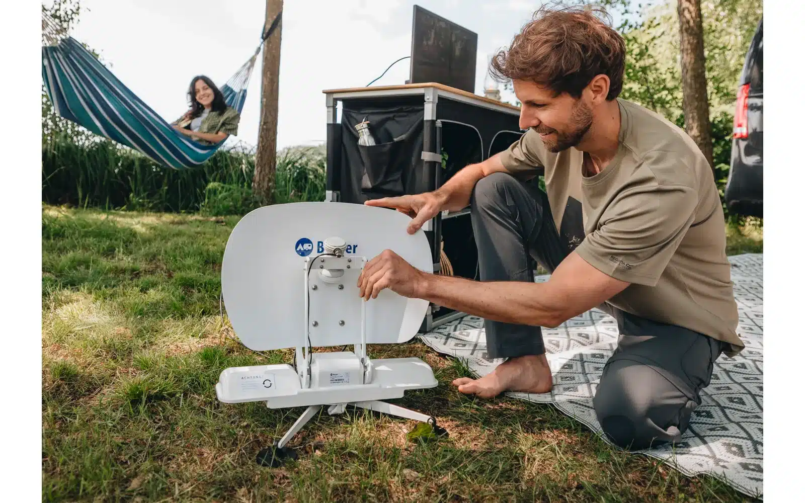 Mann richtet die Berger Pathfinder 2.0 faltbare vollautomatische Sat-Anlage im Freien ein, mit Campingausrüstung im Hintergrund.