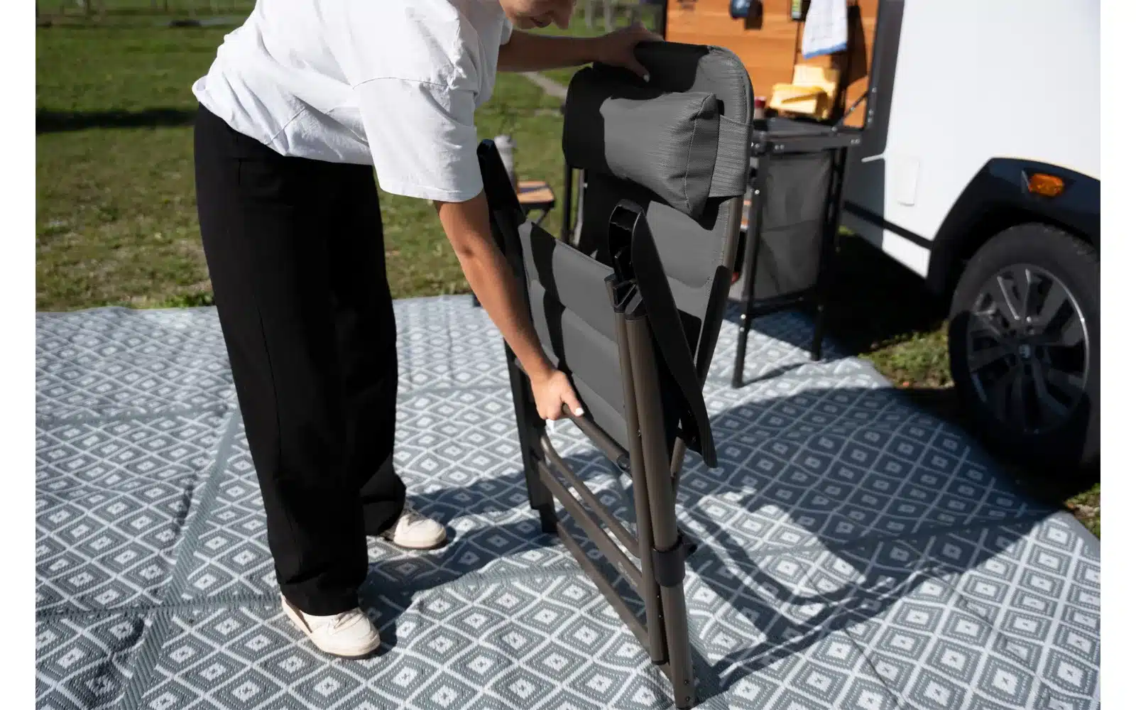 Person klappt einen Berger Slimline Klappstuhl auf einem Outdoor-Teppich vor einem Camper auf.