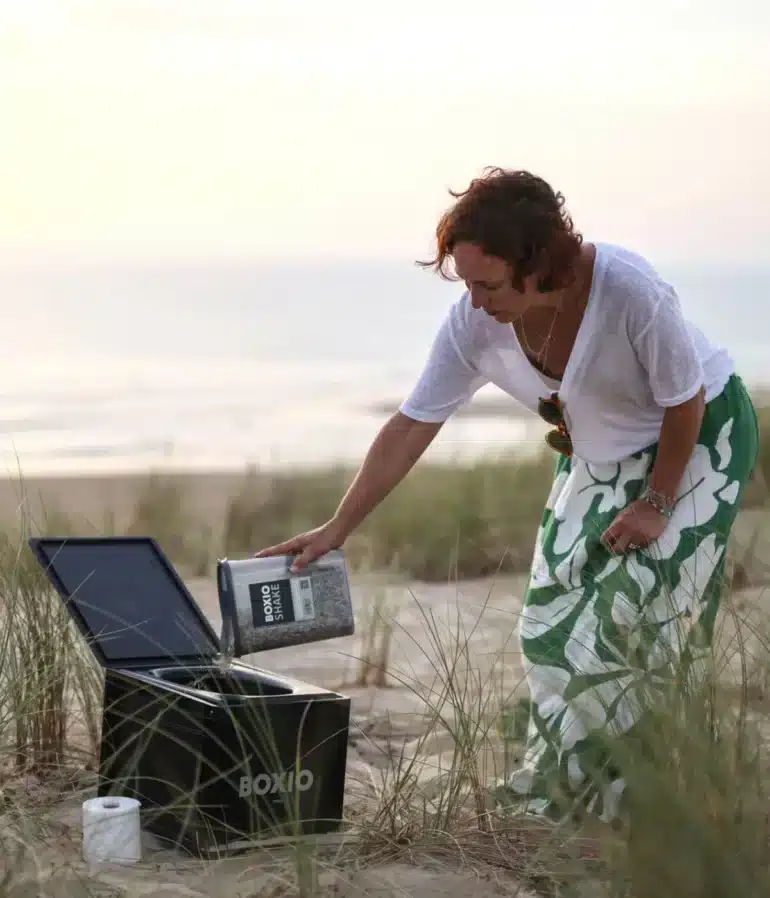 Frau befüllt eine BOXIO Trenntoilette im Freien am Strand mit Einstreu.