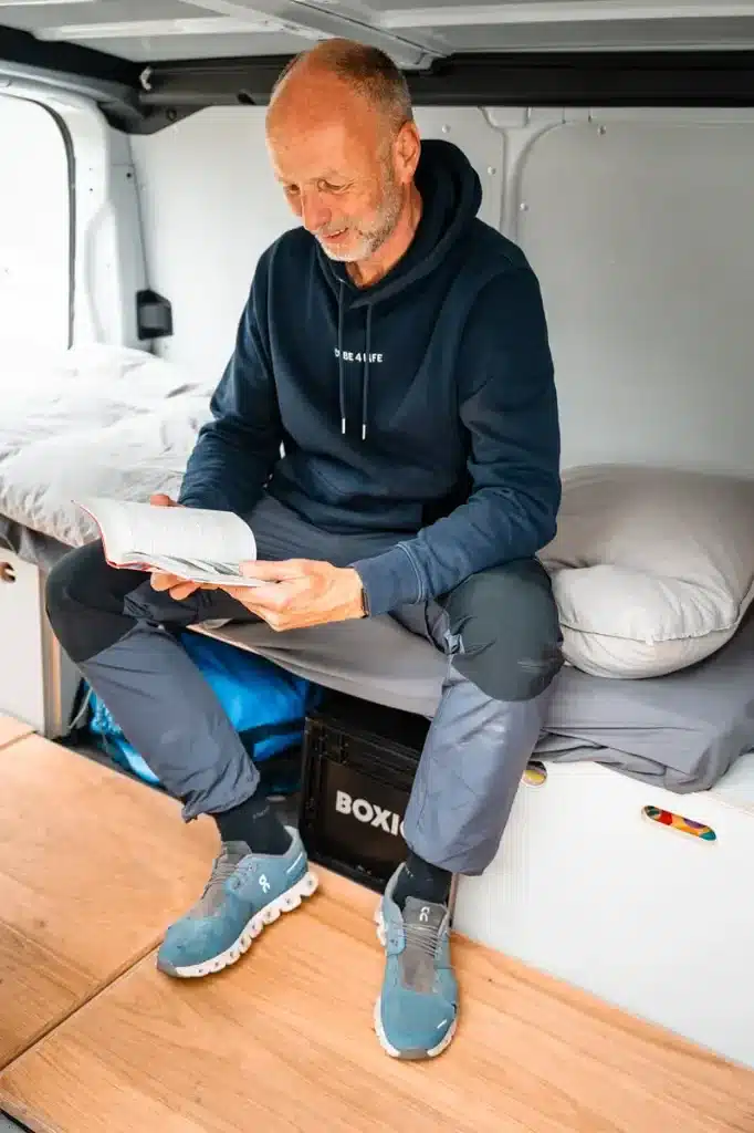 Mann sitzt im umgebauten Fahrzeug auf dem CUBE4LIFE Camper Starter Set mit Bettfunktion und liest ein Buch.