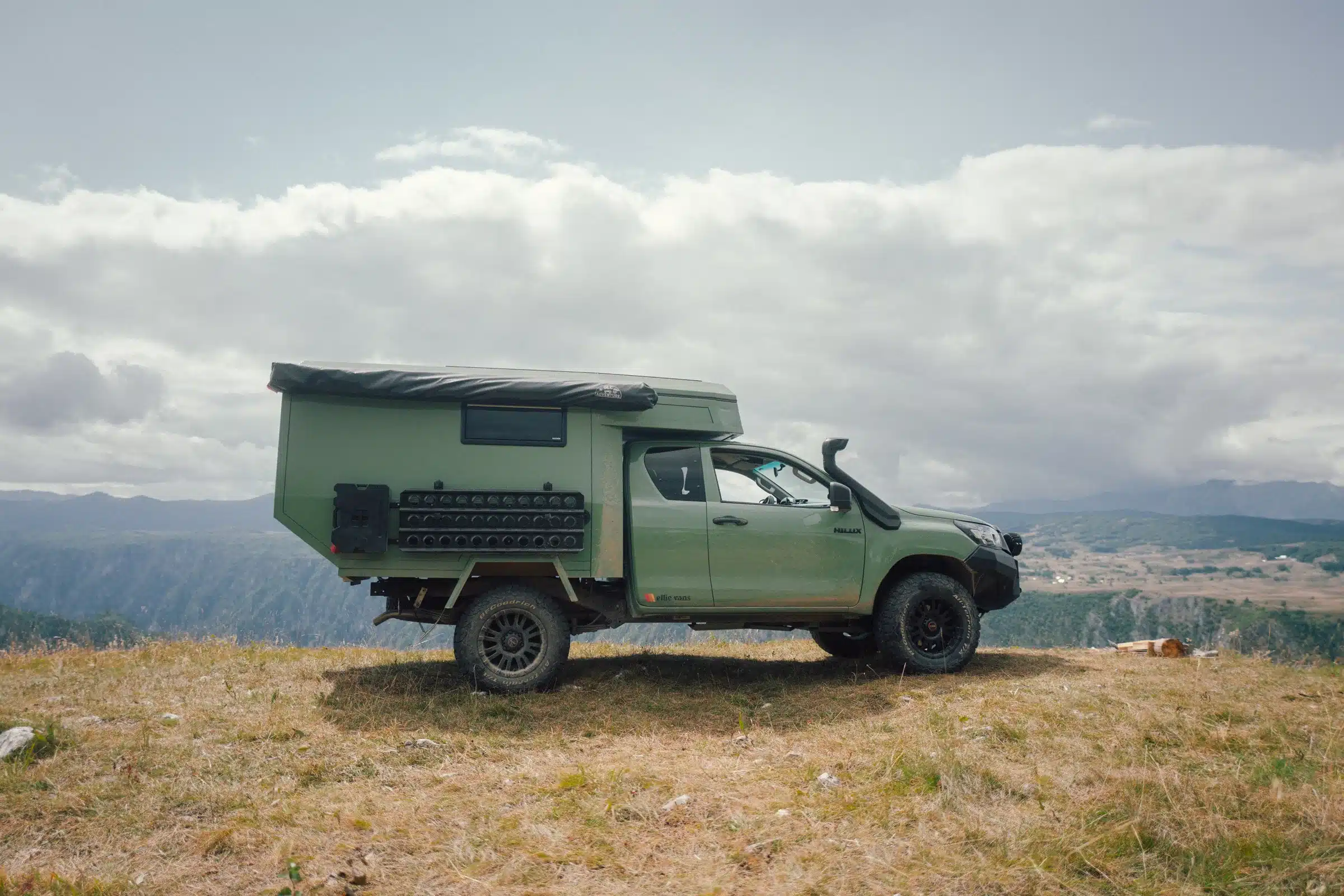 Grüner Pickup mit montierter Trailcat Expeditionskabine von Ellie Overland, stehend auf einer Wiese mit Bergen im Hintergrund