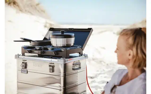 Frau sitzt am Strand vor einer Outdoor-Küche mit Campingkocher und dem mobilen Backofen VAMO von Enders.