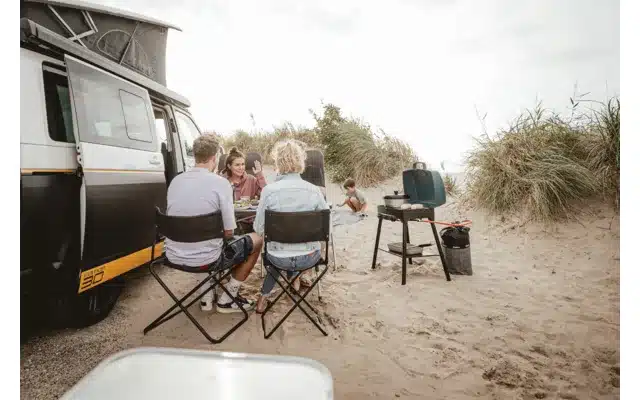 Gruppe sitzt am Strand neben einem Campingbus und Campingkocher.