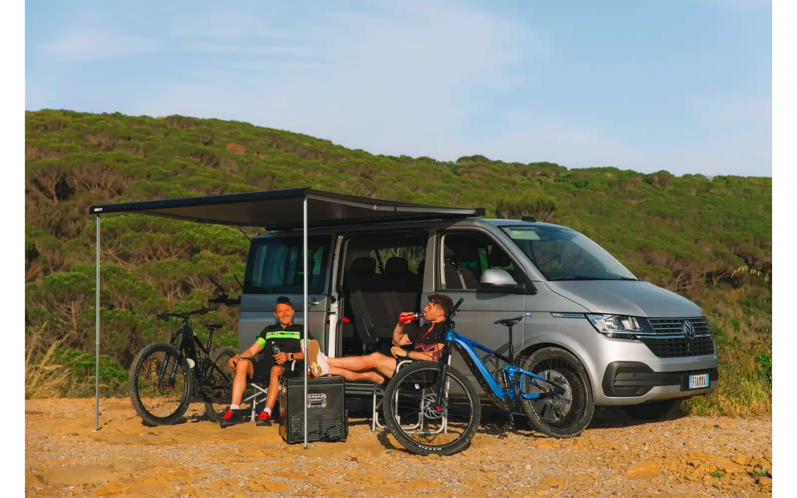 Fiamma F40 Van 270 cm Dachmarkise ausgefahren an einem silbernen VW T6 mit zwei Personen und Fahrrädern daneben auf sandigem Untergrund.