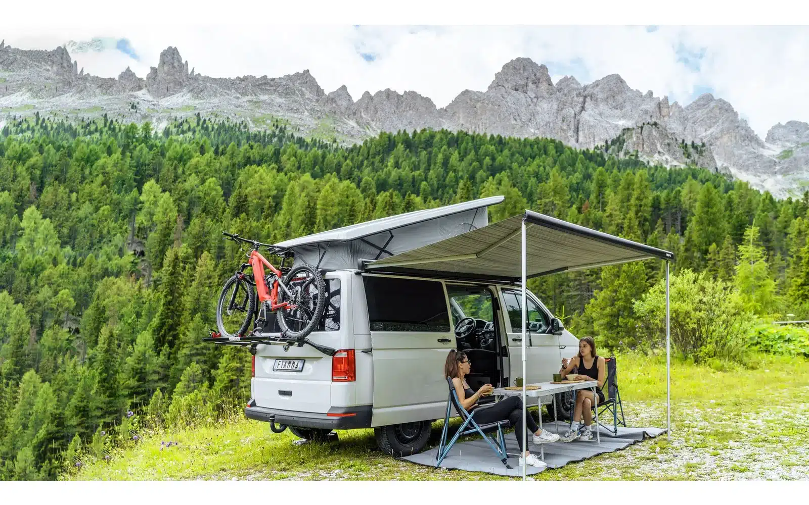 Campingbus mit ausgefahrener Fiamma F43van Markise, zwei Fahrrädern am Heck und Personen unter der Markise in bergiger Umgebung.
