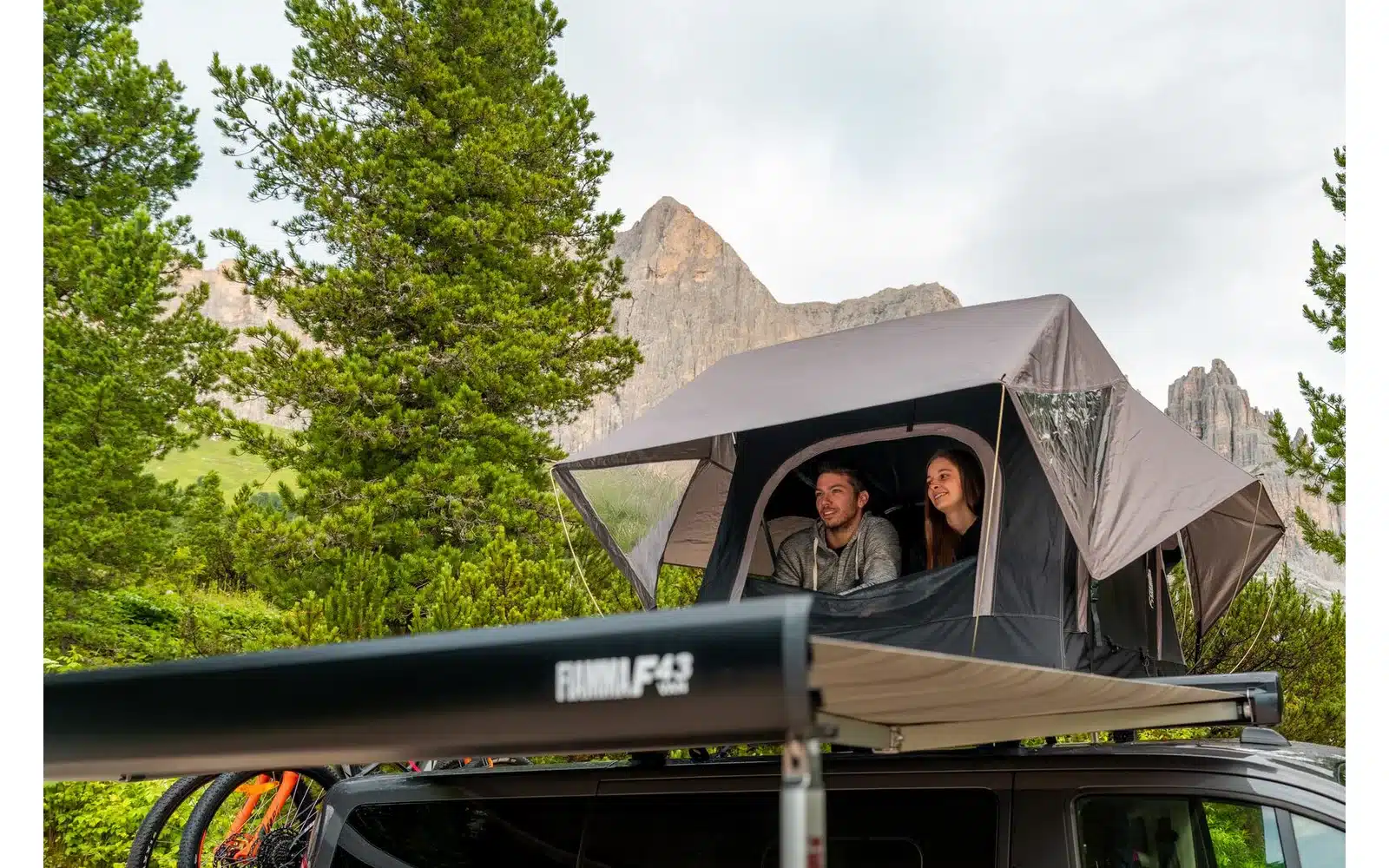 Fiamma F43van Markise an einem schwarzen Campingbus mit Dachzelt im Grünen, zwei Personen sitzen im Dachzelt, Berge im Hintergrund.