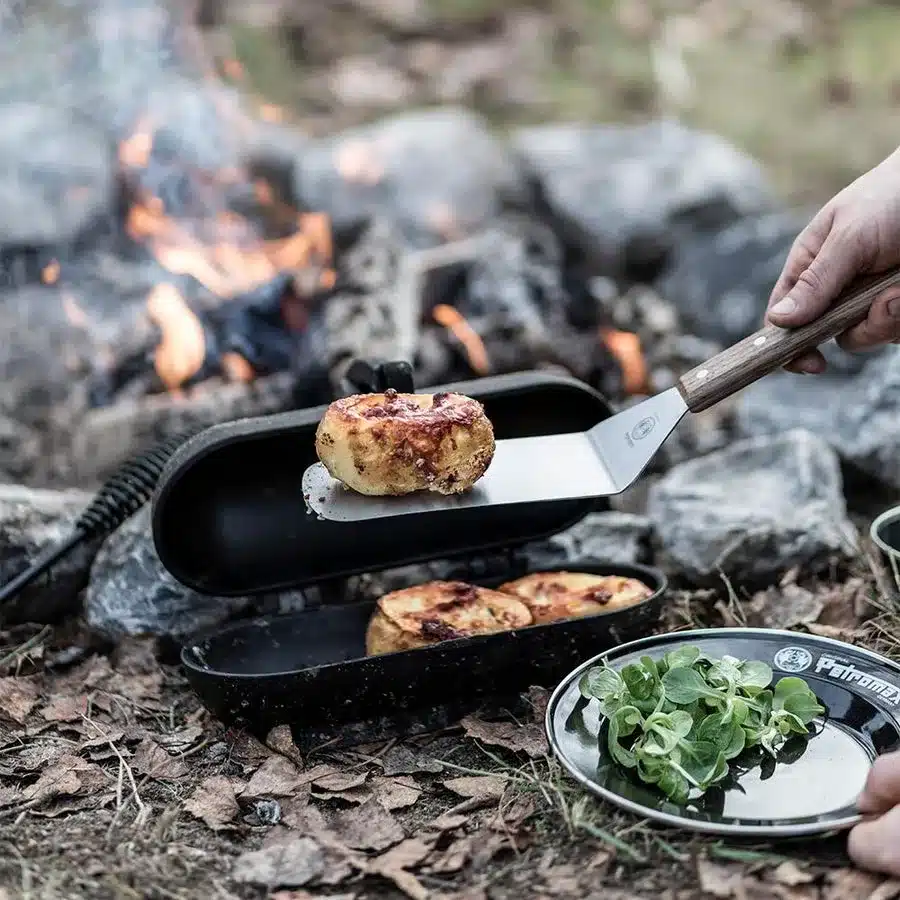 Kartoffelbräter von Petromax am Lagerfeuer mit geöffnetem Deckel, darin fertig gebratene Kartoffeln auf einem Spatenheber.