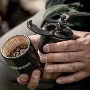 Hand hält eine Petromax Handkaffeemühle mit geöffnetem Bohnenbehälter und Gusseisen-Mahlwerk, gefüllt mit Kaffeebohnen.