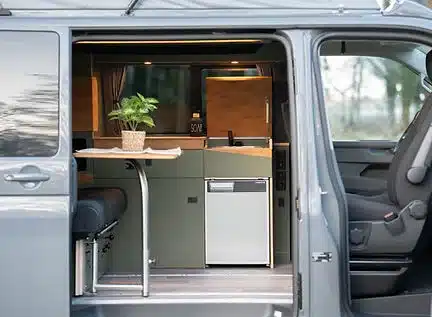Blick durch die geöffnete Schiebetür auf den Innenraum eines Vans mit eingebautem JUBO Campingausbau, sichtbarer Küchenzeile, Sitzbank und integriertem Kühlschrank.