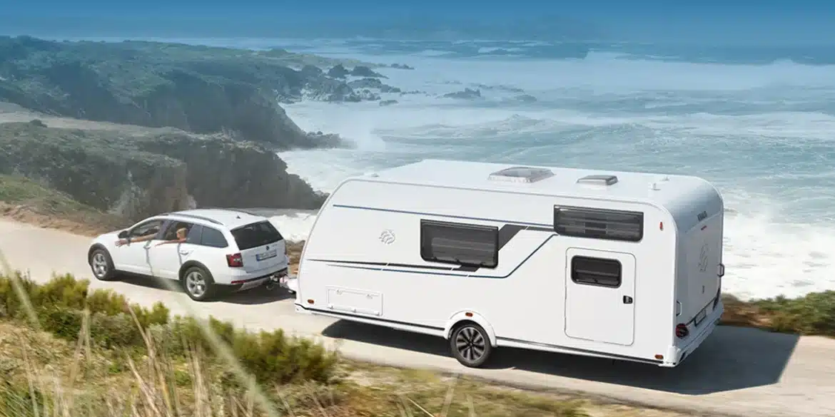 KNAUS SÜDWIND BLACK SELECTION Wohnwagen wird von einem weißen Auto entlang einer Küstenstraße gezogen.