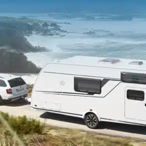 KNAUS SÜDWIND BLACK SELECTION Wohnwagen wird von einem weißen Auto entlang einer Küstenstraße gezogen.