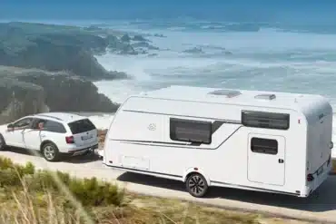 KNAUS SÜDWIND BLACK SELECTION Wohnwagen wird von einem weißen Auto entlang einer Küstenstraße gezogen.