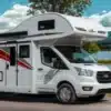 Alkoven-Reisemobil Roller Team Kronos 284 M auf einer Landstraße vor Bergkulisse und See.