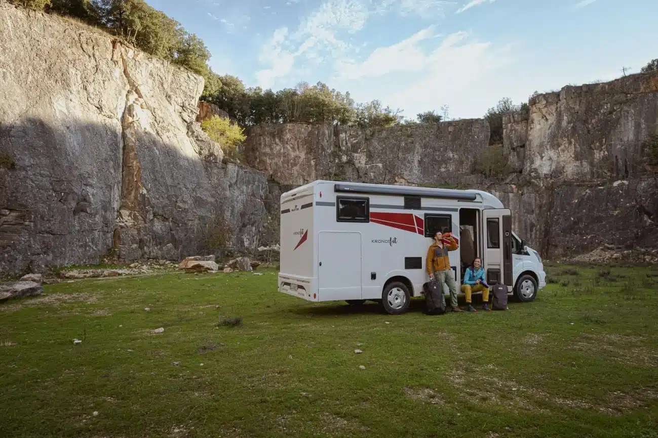 Reisemobil Kronos Fit steht auf einer Wiese vor einer Felswand, zwei Personen mit Gepäck daneben