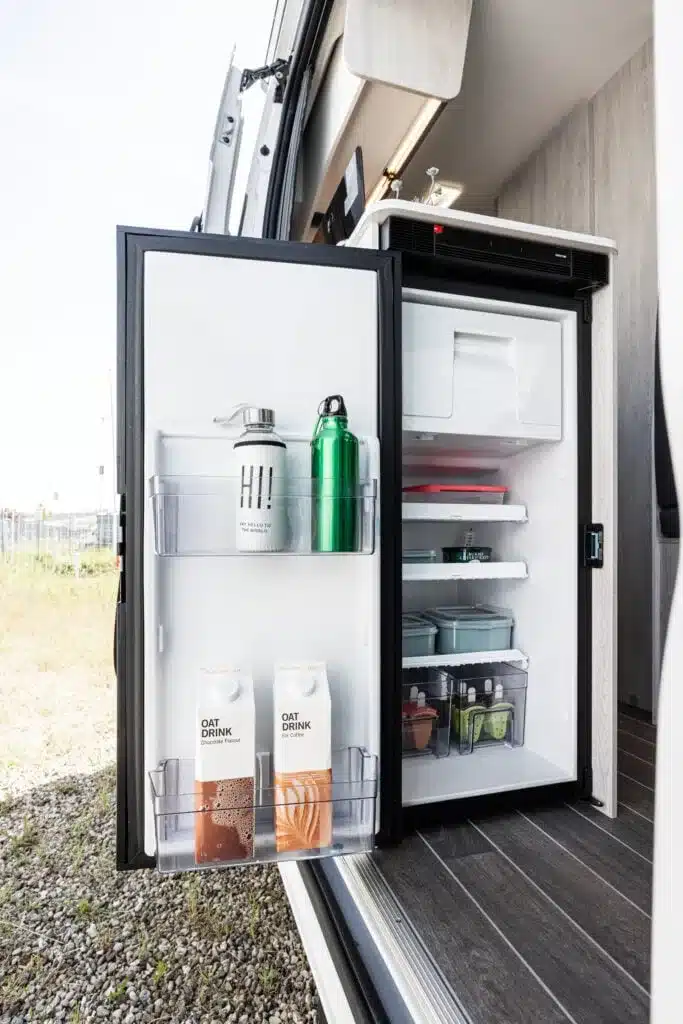 Geöffneter Kompressorkühlschrank im Innenraum des Livingstone K2 GO Campervans, gefüllt mit Getränken und Lebensmitteln.