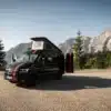 MAN TGE Next Level Campervan mit geöffnetem Aufstelldach und geöffneter Hecktür auf einer Passstraße, Berge im Hintergrund