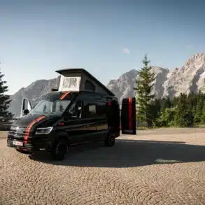MAN TGE Next Level Campervan mit geöffnetem Aufstelldach und geöffneter Hecktür auf einer Passstraße, Berge im Hintergrund