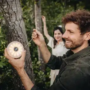 Mann befestigt die Berger 2-in-1 Campingleuchte samt Lichterkette an einem Baum, während eine Frau im Hintergrund hilft.