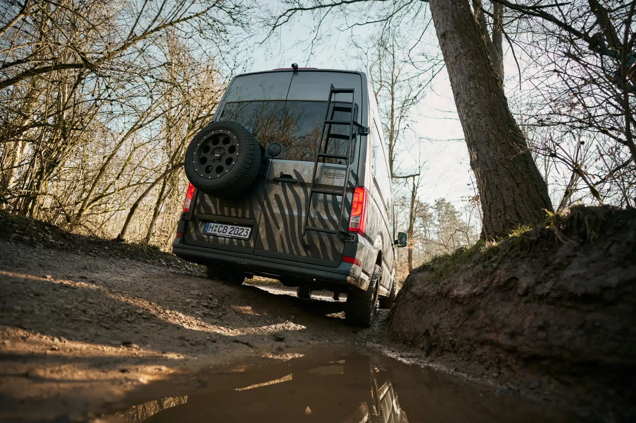 Custom-Bus Camper XL auf VW Crafter 2026, von hinten auf schlammigem Offroad-Weg mit Ersatzrad und Dachleiter, umgeben von Wald.